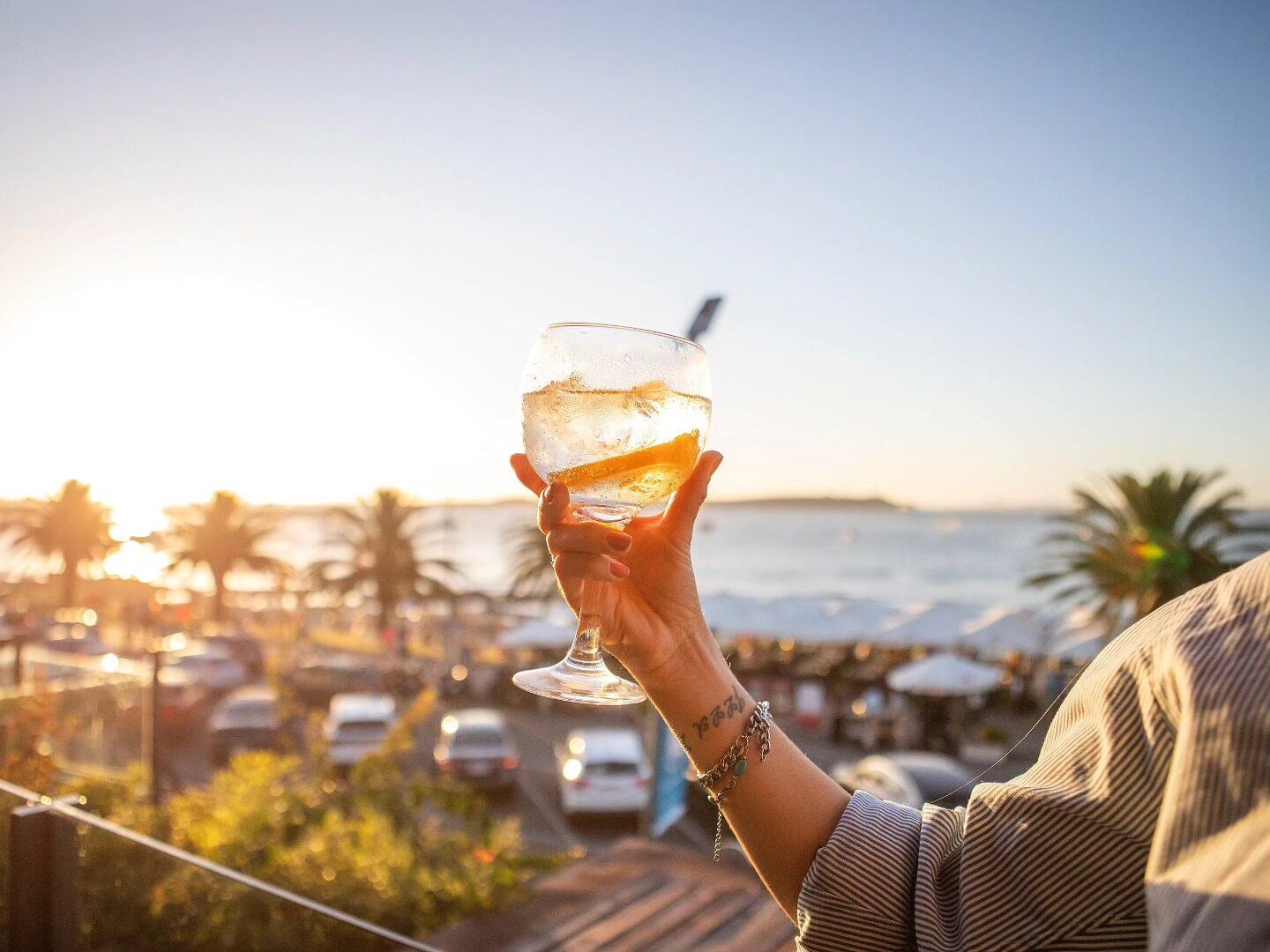 A drink held up at sunset over Playa Mansa, palm trees and the sea behind.