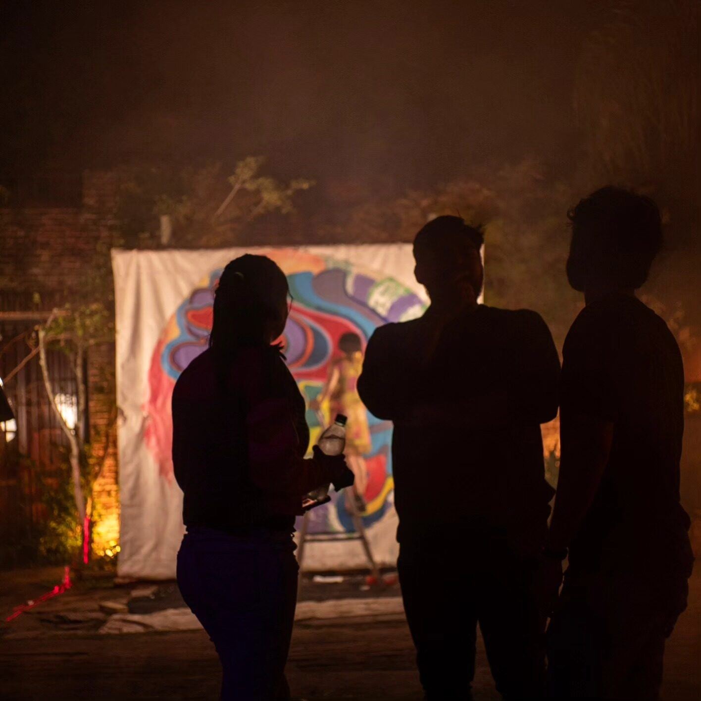 Three silhouettes facing a lit painted canvas at night.