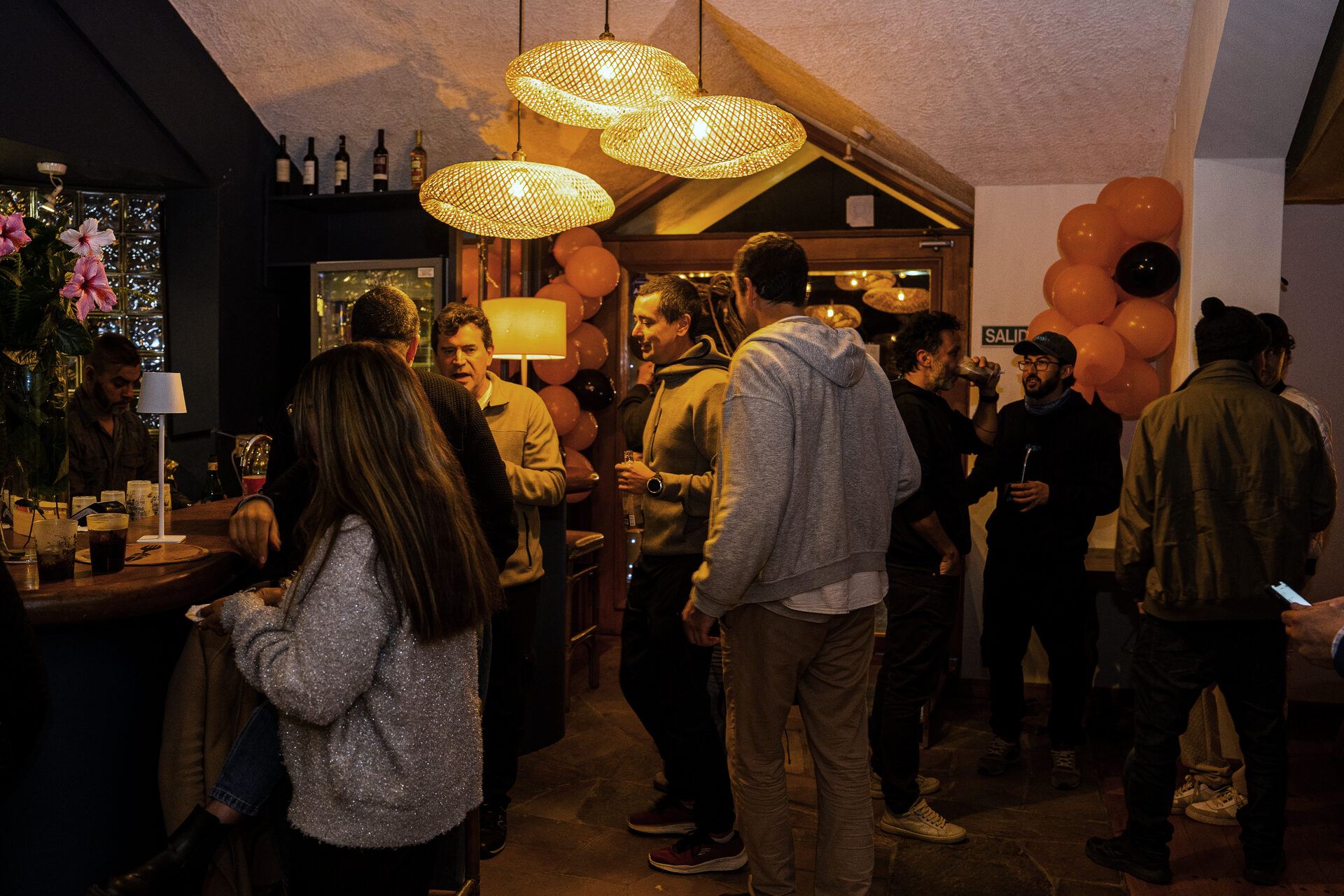 Guests talking by the bar under wicker lamps, orange balloons in the background.